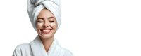 Young Caucasian woman with white towel on head and bathrobe smiling with eyes closed against white background, representing skincare and wellness.
