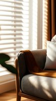 Cozy armchair with brown knitted throw blanket and white cushion near window with venetian blinds casting morning sunlight shadows in modern living room interior.