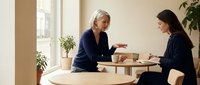 Professional mature blonde businesswoman mentoring young brunette colleague during office meeting with documents and coffee in modern workspace.