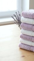 Stack of folded lavender and white towels with dried lavender sprigs on wooden surface near bright window for spa, home decor, and wellness concepts.