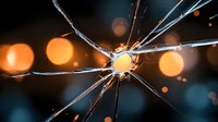 Dramatic close-up of cracked glass with radiating fracture lines against blurred orange and blue bokeh lights creating an artistic abstract pattern.