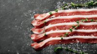 Fresh raw bacon slices arranged in row with thyme sprigs and sea salt crystals on dark stone background, food photography for restaurant menu.