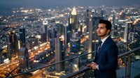 Male professional in dark business suit standing on high-rise observation deck overlooking illuminated metropolitan skyline at dusk, suitable for corporate, leadership, and business success concepts.