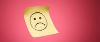 Yellow sticky note with hand-drawn sad face emoticon on pink background for office communication, workplace feedback, and emotional expression concepts.