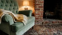 Cozy living room interior with green velvet armchair, fur throw, warm lamp lighting and exposed brick fireplace creating comfortable home atmosphere.
