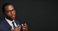African American male professional in navy blazer and tie wearing eyeglasses, gesturing while speaking against dark background for corporate communication and business presentation concepts.