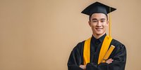 Young Asian male graduate in black cap and gown with yellow stole, arms crossed, smiling confidently against neutral beige background.