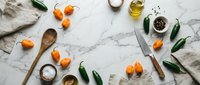 Fresh orange kumquats with green leaves scattered on white marble surface alongside wooden spoons, olive oil, salt and linen napkins for culinary preparation.