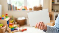 Child holding blank white card in playroom filled with colorful toys and wooden storage, ideal for educational marketing and childcare advertising.