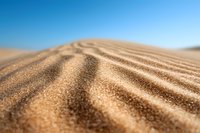 Close-up macro view of sand dunes with rippled texture against clear blue sky background, natural desert landscape pattern showing wind-carved formations.