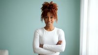 Young African American businesswoman with curly hair in white shirt standing confidently with crossed arms in modern office space.