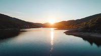 Tranquil mountain lake at sunset with golden sunlight reflecting on calm water surface, surrounded by rolling hills creating serene wilderness landscape.