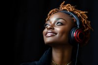 Young African American woman with natural curly hair wearing headphones smiling against dark background, expressing joy while listening to music.