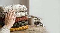 Woman's hand touching stack of cozy knitted sweaters in warm autumn colors on wooden surface, perfect for seasonal wardrobe and home comfort concepts.