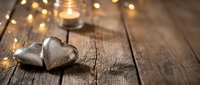Two rustic metal hearts on weathered wood planks with warm golden bokeh lights and glowing candle creating romantic Valentine's Day atmosphere.