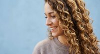 Young Caucasian woman with curly blonde hair smiling softly, wearing a cozy sweater against a light blue background.