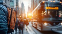 Young commuter with brown leather backpack walking in busy city street at sunset with bus and crowd, urban transportation scene with golden light flare.