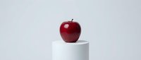 Red apple sitting on white cylindrical pedestal against clean gray background for minimalist food photography and healthy eating concepts.