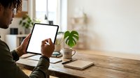 Young man with dark skin tone working on tablet device at wooden desk with coffee cup and notebook in bright home office space.