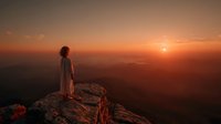 Woman in flowing dress standing on mountain cliff edge watching golden sunrise over layered mountain ranges creating peaceful meditation moment.