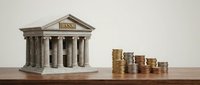 Miniature bank building with classical columns next to stacks of golden coins on wooden surface representing financial growth and banking services.