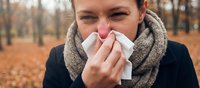Woman blowing nose with tissue in autumn park, suffering from seasonal cold, flu or allergies. Healthcare concept with fallen leaves background.