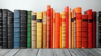 Row of colorful vintage hardcover books arranged by color spectrum from dark blue to orange and red on wooden shelf against gray wall.