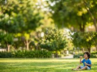 Young boy in blue shirt sitting on grass in sunny park, blowing bubbles outdoors during daytime recreational activity.