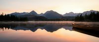 Wooden boat rests on calm lake at sunrise with snow-capped mountain peaks reflected in still water, surrounded by evergreen forest during golden hour.