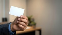 Young adult male with dark skin tone wearing business casual attire holding blank white card in office environment for corporate branding and professional networking purposes.