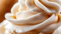 Creamy white soft serve ice cream or whipped cream swirls in macro close-up showing smooth texture and delicate peaks against warm background.