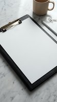 Black leather clipboard with blank white paper, silver pen and beige coffee mug on marble desk surface for business planning and office productivity.