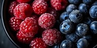 Fresh ripe raspberries and blueberries with water drops in dark bowl, macro shot of mixed berries. Natural organic summer fruits for healthy eating.