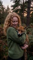 Young Caucasian woman with curly blonde hair holding red fox in forest during golden hour sunset creating warm magical wildlife connection moment.