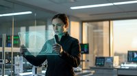 Young professional woman with light skin tone wearing black business attire holding glowing digital shield interface in modern office environment, representing cybersecurity, data protection, and technology innovation.