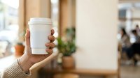 Male hand holding blank white takeaway coffee cup in modern cafe interior with blurred background, suitable for beverage branding, coffee shop marketing, and hospitality advertising.