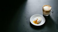 Soft-boiled egg with runny yolk on white plate next to cappuccino in glass cup on dark surface, overhead view for breakfast and food styling projects.