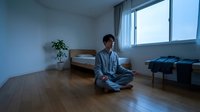 Young Asian man in pajamas meditating in lotus position on wooden floor in minimalist bedroom with natural light from large window.