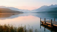 Serene mountain lake at sunrise with wooden dock and distant kayaker, peaceful morning reflection creating tranquil landscape for relaxation and outdoor recreation.