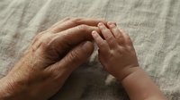 Elderly grandmother hands gently holding baby hands showing intergenerational love and family bond on soft textile background.