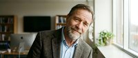 Mature male professional with gray beard wearing casual blazer and light blue shirt in modern office environment with natural window light.