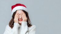 Young woman in Santa hat covering face with hands, wearing white sweater against gray background. Perfect for holiday stress concepts.