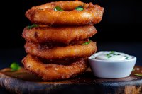 Crispy golden onion rings stacked on rustic wooden board with creamy dip sauce and fresh herbs. Dark moody background highlights appetizing texture.
