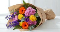 Colorful mixed flower bouquet wrapped in kraft paper with orange gerberas, yellow roses, purple chrysanthemums and greenery on white surface.