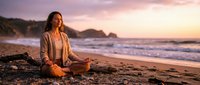 Young Caucasian woman meditating in lotus pose on sandy beach at golden sunset, ocean waves and cliffs in warm background.