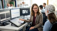 Professional businesswoman with glasses presenting data analytics on dual monitors to colleagues in modern office workspace environment.