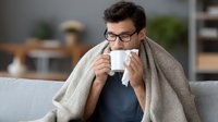 Sick Caucasian man wrapped in blanket drinking hot tea while sitting on couch at home, treating cold and flu symptoms during winter season.