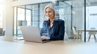 Confident mature businesswoman with silver hair working on laptop in modern glass office space, professional corporate environment for business success.