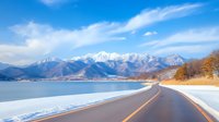 Scenic winter road along frozen lake with snow-capped mountains and wispy clouds in blue sky, curving asphalt highway leads to distant peaks.