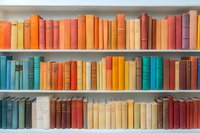 Colorful vintage books arranged on white wooden shelves, spines in rainbow order from orange and red to turquoise and blue, creating organized library display.